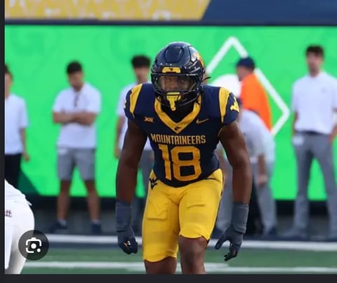 Football player wearing Mountaineers #18 jersey in black and gold uniform stands on field with coaches in background.