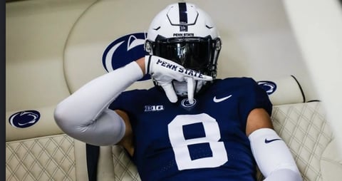 Penn State football player wearing number 8 navy jersey adjusting white helmet in locker room